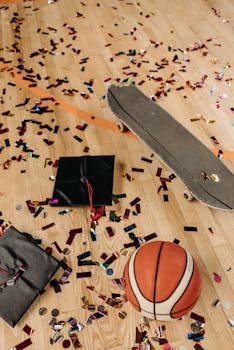 A graduation cap, basketball, and skateboard on a confetti-covered hardwood floor.