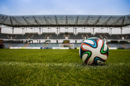 the-ball-stadion-football-the-pitch-46798-46798 Close-up of a soccer ball on a lush grass field with an empty stadium in the background.